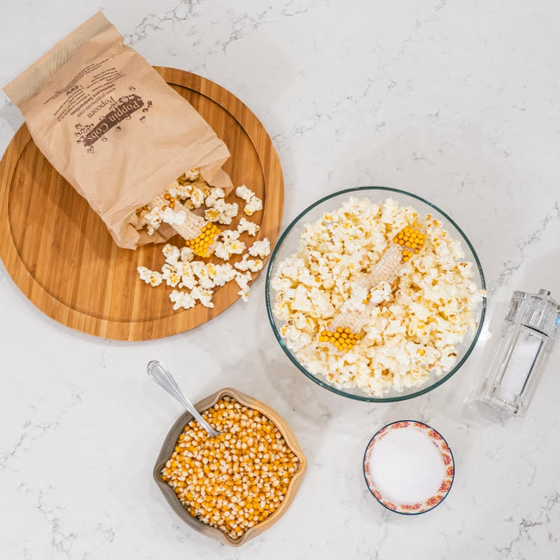 An overhead photograph of a Poppin' Cob popcorn making setup, including a wooden board with popcorn, a bowl of popcorn kernels, seasonings, a bulk bag of popcorn, and printed instructions for popping.