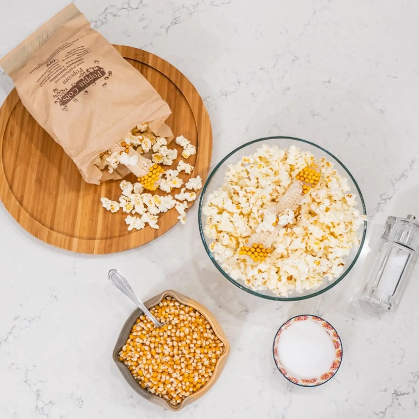 Popcorn-making setup with a bag of popcorn kernels, a bowl of popped popcorn, and a container of unpopped kernels on a marble surface.